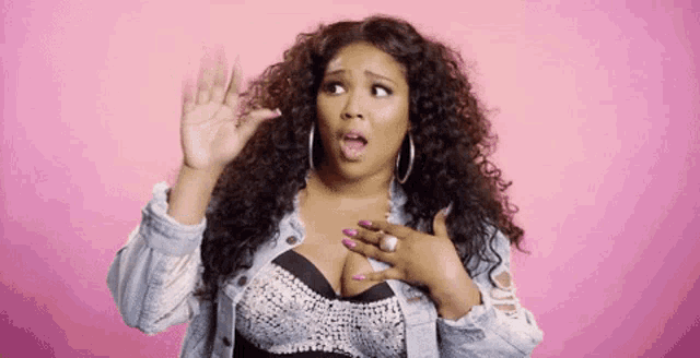 a woman with curly hair and a denim jacket is making a funny face on a pink background .