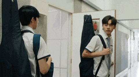 two young men are standing next to each other in a hallway . one of the men is holding a guitar case .