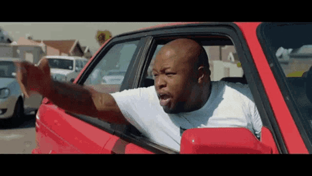 a man in a white shirt is driving a red car and giving the middle finger