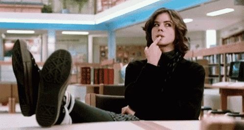 a woman is sitting at a table in a library with her feet up and her finger on her chin .