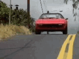 a red car is driving down a road next to a grassy hill .