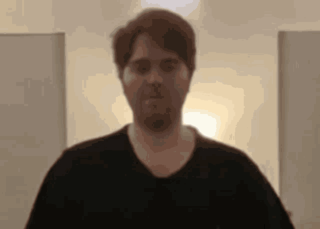 a man with a beard is wearing a black shirt and standing in a room with a light on .