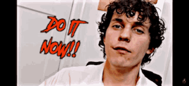 a man with curly hair is standing in front of a wall that says do it now