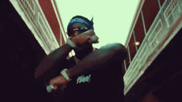 a man wearing sunglasses and a bandana is standing in front of a building and singing into a microphone .