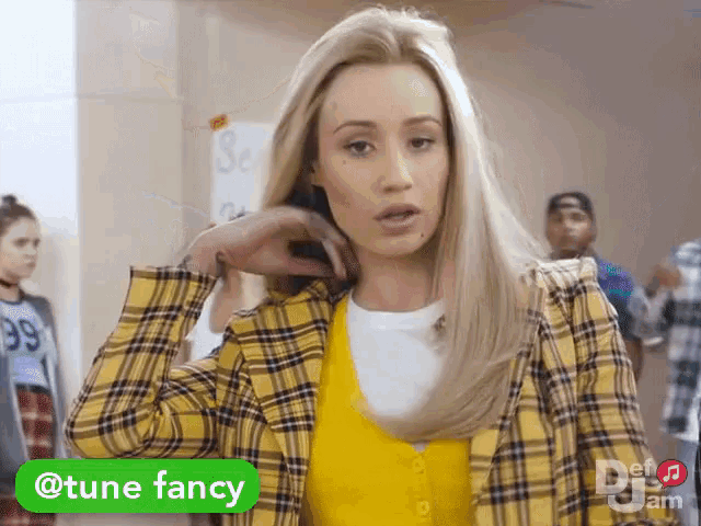 a woman wearing a plaid jacket and a yellow vest has a green sticker that says tune fancy on it