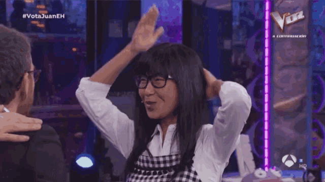 a woman wearing glasses and a plaid shirt is raising her hands in the air in front of a sign that says voz