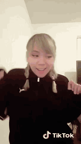 a woman in a black jacket is dancing in a room with a tiktok logo on the bottom .