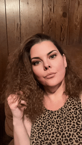 a woman in a leopard print top holds her hair in front of a wooden wall