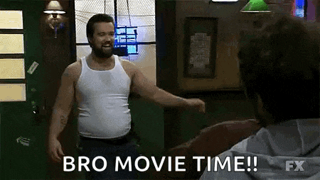 a man in a white tank top is standing next to another man in a room and says `` bro movie time ! ''