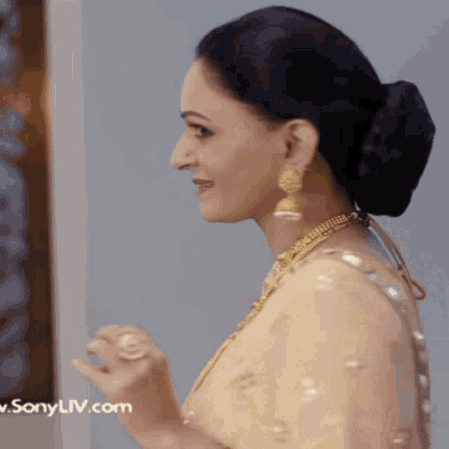 a woman wearing a gold necklace and earrings is standing in front of a sonyliv.com logo