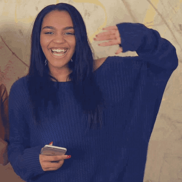 a woman in a blue sweater holds a cell phone and smiles