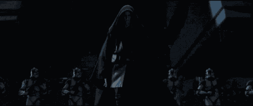 a man in a hooded cloak is standing in front of a group of stormtroopers in a dark room .