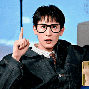 a young man wearing glasses and a denim jacket points upwards