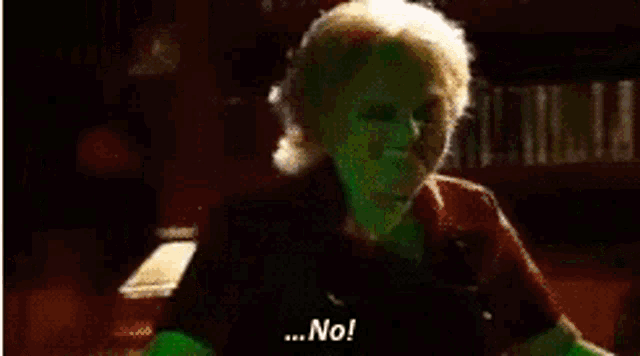 a woman with a green face is standing in front of a bookshelf and says `` no '' .