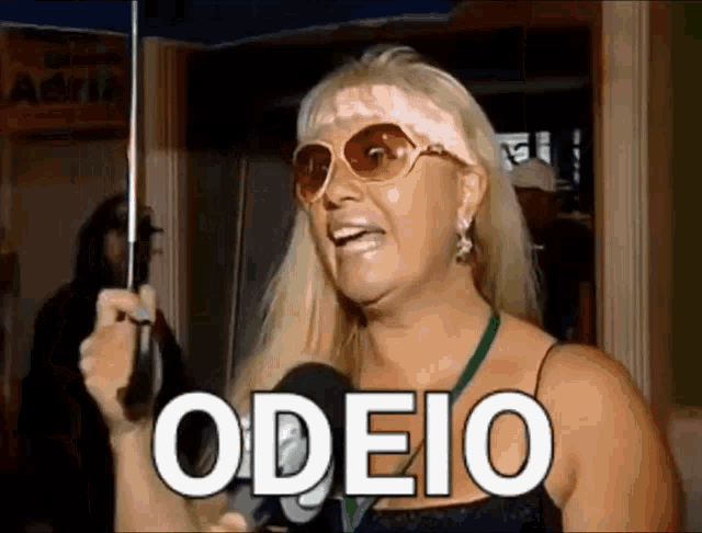 a woman wearing sunglasses and holding an umbrella is talking into a microphone and says odeio .