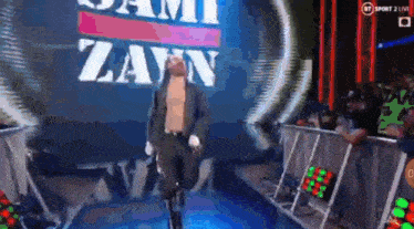 a man in a suit is walking into a wrestling ring with the word zayn on the screen behind him .