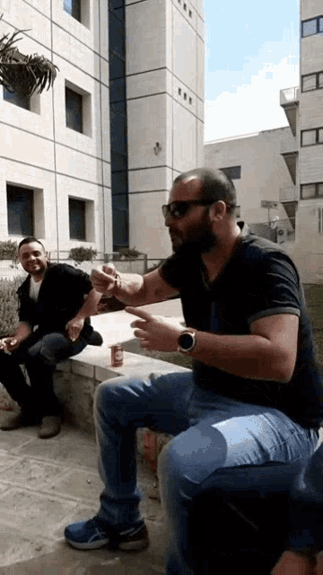 two men are sitting on a sidewalk in front of a building and one is wearing sunglasses