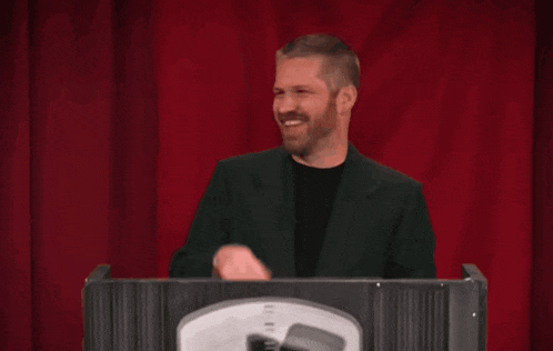 a man with a beard is sitting at a podium with a red curtain behind him