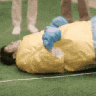a person is laying on a green field wearing a yellow shirt and blue gloves .
