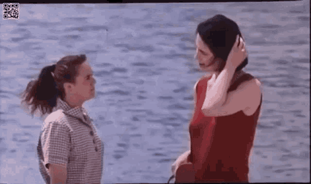 two women are standing next to each other in front of the water and talking to each other .