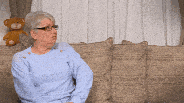 an older woman wearing glasses sits on a couch with a teddy bear in the background
