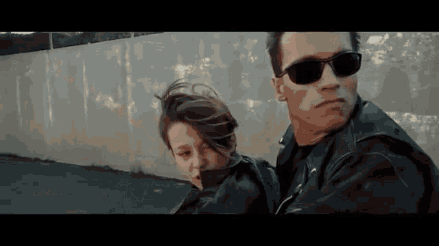 a man and a woman are standing next to each other in front of a wall wearing sunglasses .
