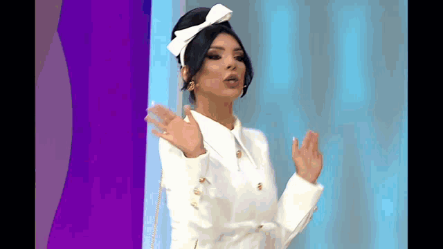 a woman wearing a white jacket and a white bow in her hair is standing in front of a blue curtain .