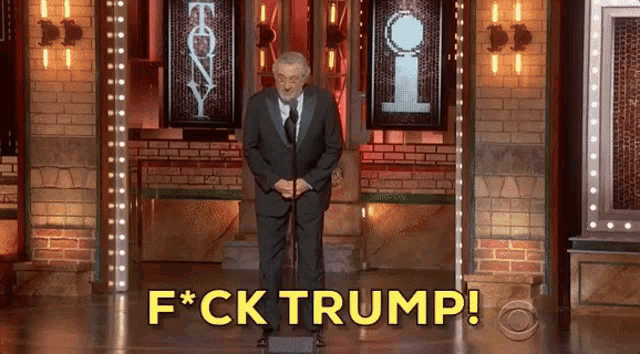 a man in a suit and tie stands in front of a microphone with the words f * ck trump above him
