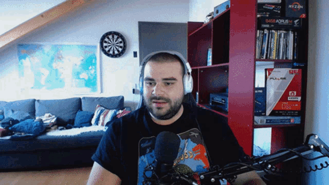 a man wearing headphones stands in front of a microphone in a living room with a dart board on the wall above him