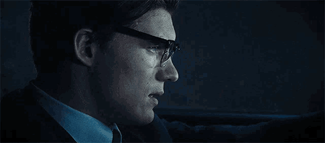 a man wearing glasses and a suit is driving a car .