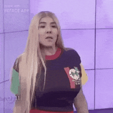 a woman with long blonde hair is standing in front of a purple wall wearing a blue shirt .