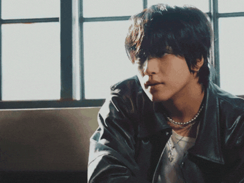 a man wearing a pearl necklace and a leather jacket is sitting in front of a window