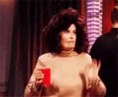 a woman in a tan sweater is holding a red cup and waving .