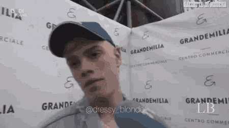 a man wearing a hat is talking into a microphone while standing in front of a banner that says grandemilia .