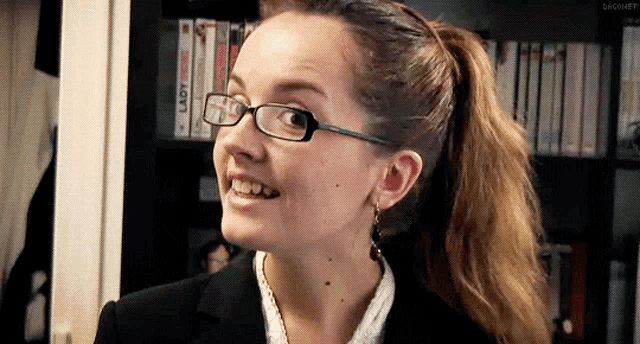 a woman wearing glasses and a ponytail is smiling in front of a shelf with lady dvds on it