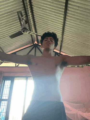 a shirtless man stands in front of a ceiling fan with his arms outstretched