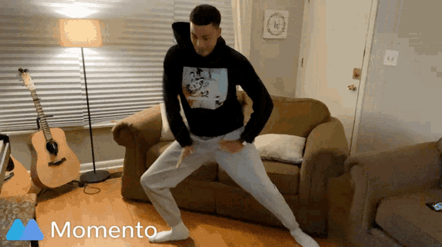 a man in a black hoodie is dancing in a living room with a momento logo in the corner