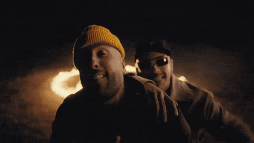 a man wearing a yellow beanie and sunglasses stands next to another man