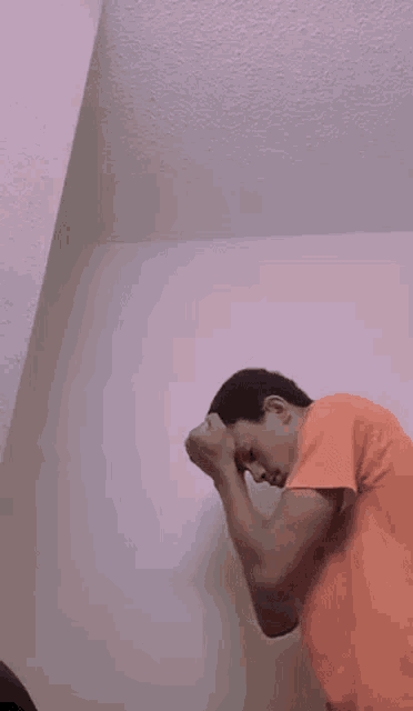 a man in an orange shirt is leaning against a wall with his hands on his face .