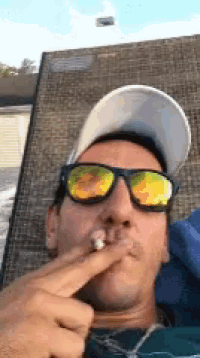 a man wearing sunglasses and a hat is smoking a cigarette while sitting in a chair .
