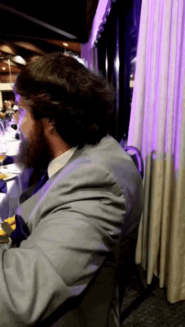 a man in a suit sits at a table with a purple curtain behind him
