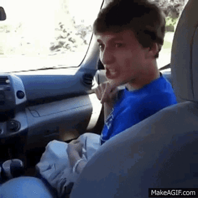 a young man in a blue shirt is sitting in the driver 's seat of a car