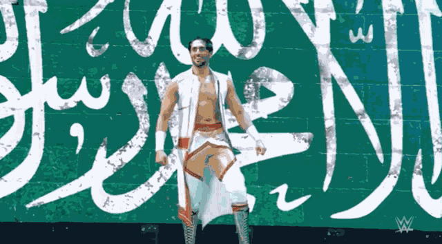 a wrestler stands in front of a green wall with arabic writing on it