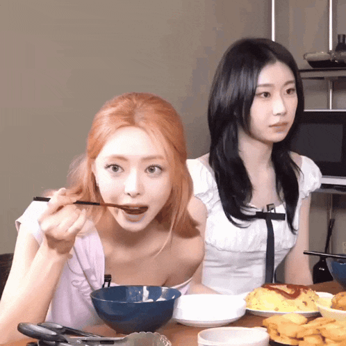two women are sitting at a table with plates of food and one is eating with chopsticks
