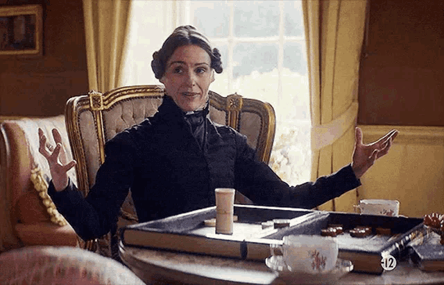 a woman in a black dress is sitting at a table with a tray of cookies and tea cups .