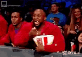 a man in a red jacket is eating popcorn while watching a wrestling match .