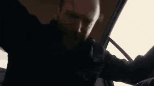a man with a beard is sitting in the driver 's seat of a car with his arms outstretched .