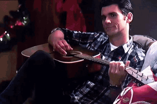a man in a plaid shirt sits on a couch playing an acoustic guitar
