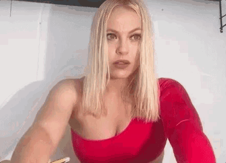 a blonde woman is wearing a red one shoulder top and taking a selfie .