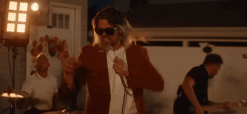 a man wearing sunglasses is singing into a microphone in front of a band